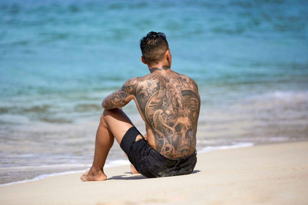 a man sitting on a beach with a heavily tattooed back