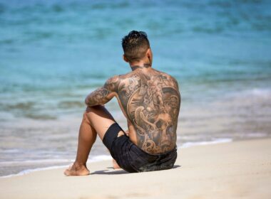 a man sitting on a beach with a heavily tattooed back