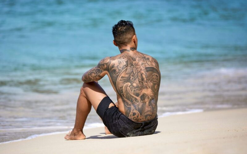 a man sitting on a beach with a heavily tattooed back
